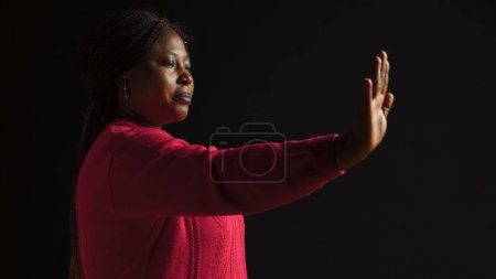 Photo for African american woman firmly signals stop using hand gesture in isolated black background. Female fashion model expresses disapproval rejecting and disagreeing in side-view portrait shot. - Royalty Free Image