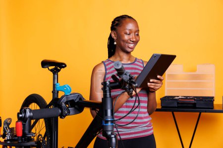 Photo for Dedicated young woman repairing a broken bicycle with tools and technology, against an isolated yellow background. Female african american cyclist thoroughly inspecting bicycle with digital tablet. - Royalty Free Image
