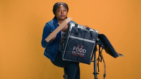 Mujer negra en bicicleta entregando cuidadosamente comida fresca para llevar con bolsa de mensajería. Persona africana americana de la entrega que proporciona servicio al cliente profesional haciendo entrega eficiente.