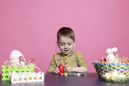 Foto de Adorable jovencito haciendo arreglos encantadores para el Domingo de Pascua y pintando huevos con sellos y pinceles. Delicioso niño le gusta usar materiales de elaboración para decorar en el estudio. - Imagen libre de derechos