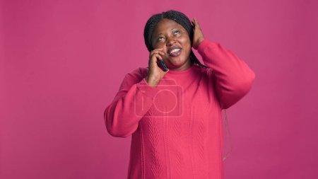 Vibrant young black woman moving around and happily speaking on mobile phone. Female african american beauty in front of isloated background having funny conversation through cell phone device.