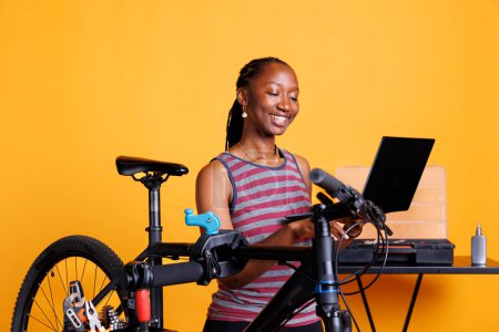 Energetische schwarze Frau inspiziert und repariert Fahrrad, nutzt Laptop und spezialisiertes Werkzeugset für Wartung und Reparatur. Sportliche afrikanisch-amerikanische Dame mit Minicomputer repariert Fahrradschäden.