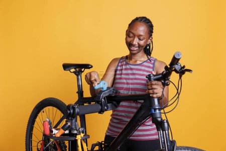 Energetische schwarze Frau, die Anpassungen vornimmt und Fahrräder mit speziellen Werkzeugen repariert. Engagierte Radfahrerin greift und begutachtet Fahrradrahmen auf Reparaturständer vor gelbem Hintergrund.