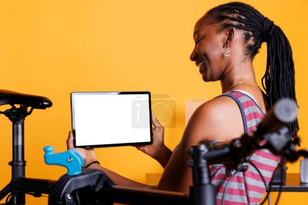 Photo for Sports-loving black woman stands by broken bicycle while carrying tablet with blank chromakey mockup template horizontally. African american female grasps digital gadget that displays a whitescreen. - Royalty Free Image