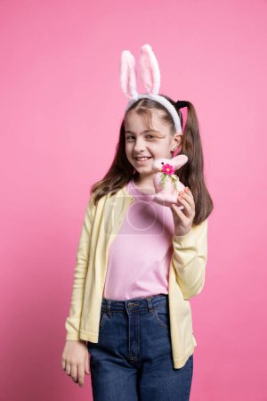 Foto de Adorable niña pequeña sosteniendo un juguete de conejo rosa esponjoso en la cámara, presentando sus juguetes y adornos coloridos de Pascua. Joven niño feliz con orejas de conejo sintiéndose alegre y emocionado por la festividad. - Imagen libre de derechos