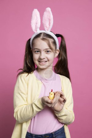Foto de Niña sonriente sosteniendo a una adorable chica de peluche delante de la cámara, un niño alegre con un juguete dorado esponjoso sobre un fondo rosa. Niño pequeño con orejas de conejito lindo para la festividad. - Imagen libre de derechos