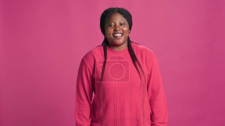Atemberaubende afrikanisch-amerikanische Dame in rosa stylischem Outfit lacht für die Kamera beim Fotoshooting. Lebendiges weibliches Fashionista-Model, das vor rosa Hintergrund gluckst. Zeitlupe, Porträtaufnahme.