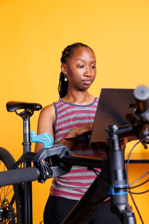 Ciclista afroamericana que utiliza tecnología digital para el mantenimiento y reparación de bicicletas rotas. Vista detallada de la mujer negra deportiva que navega en el ordenador portátil para instrucciones de mantenimiento de bicicletas.