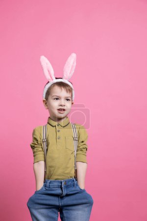 Téléchargez les photos : Jeune garçon adorable souriant et debout sur fond rose, portant des oreilles de lapin pour célébrer la fête de Pâques. Petit enfant joyeux étant heureux devant la caméra, attendant de recevoir des cadeaux. - en image libre de droit