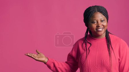 Photo for Glamorous african american fashionista confidently poses her stylish pink sweater outfit against vibrant backdrop. Young black woman with elegant style motioning to her right-side with her hand. - Royalty Free Image