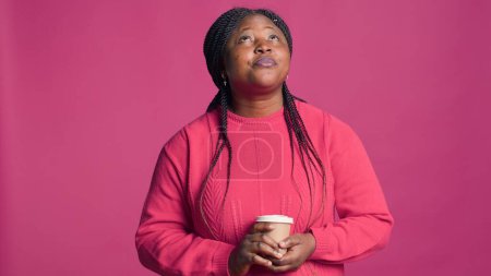 Porträt einer Afroamerikanerin, die in Gedanken aufblickt und eine Tasse Kaffee in der Hand hält. Zufriedene Modebloggerin im rosafarbenen Pullover genießt Heißgetränk.