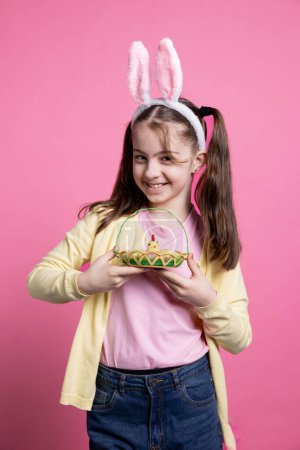 Photo for Lovely little kid with rabbit ears poses with adorable springtime toys while carrying an Easter basket full of eggs and a chick. Joyful and enthusiastic youngster enjoying the holidays in studio. - Royalty Free Image