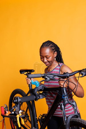 Aktives gesundes Damenfahrrad auf Reparaturständer zur Reparatur vor gelbem Hintergrund. Bild zeigt eine sportliche schwarze Frau, die ihre Fahrradgröße anpasst, bereit für die jährliche Wartung.