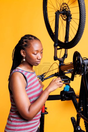 Deportiva mujer afroamericana ciclista reparando y manteniendo para asegurar un rendimiento de primera clase. Vista detallada de la mujer negra sana examinando meticulosamente y corrigiendo el pedal de la bicicleta.