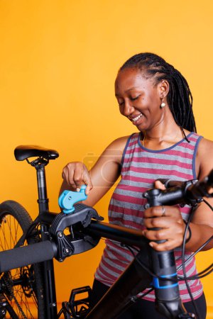 Eine afroamerikanische Radfahrerin inspiziert ihren kaputten Fahrradrahmen auf einem Reparaturständer vor isoliertem gelben Hintergrund. Schwarze Frau bereitet sich auf Anpassungen und Reparaturen mit Spezialgeräten vor.
