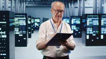 Senior technician looking around high tech data center, using clipboard to crosscheck disaster recovery plan and assess server parts in need of replacement, preventing damages