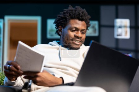 Photo for Joyful freelancer working from apartment, writing down business ideas while watching online video. Smiling man using pen and paper to note strategy plan, learning from internet clip on laptop - Royalty Free Image