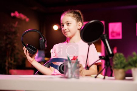 Youngster presents headphones in different price ranges to audience, comparing them to find winner, filming in neon lit home studio. Gen Z tech influencer child recommends listening devices to viewers