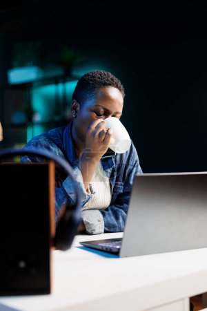 Foto de Retrato de una trabajadora remota afroamericana tomando café mientras trabajaba en su portátil digital. Joven mujer negra con computadora inalámbrica en el escritorio está bebiendo de una taza. - Imagen libre de derechos