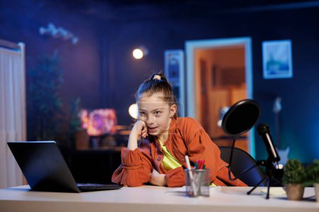 Kleine Mädchenfilme mit professioneller Ausrüstung in blauem neonbeleuchtetem Wohnzimmer, das als Vlogging-Studio dient. Junge Influencerin filmt mit High-Tech-Kamera und diskutiert über Themen, die bei Kindern beliebt sind