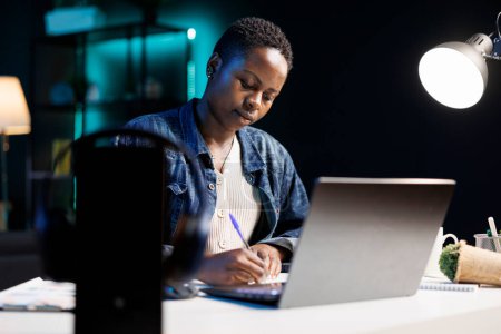 Foto de La mujer afroamericana trabaja diligentemente en su escritorio usando su computadora portátil y su pluma, investigando e ideando ideas. Un individuo productivo y enfocado que utiliza la tecnología para el trabajo y el estudio. - Imagen libre de derechos