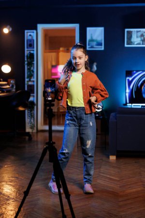 Girl dancing and singing in personal studio while shooting video with cellphone on tripod for social media. Joyful kid entertains subscribers with professional choreography recorded with mobile phone