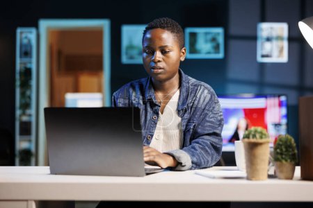 Photo for Busy black female freelancer sitting in her modern living room office, working on personal computer. African american college student typing and reviewing information on digital laptop. - Royalty Free Image