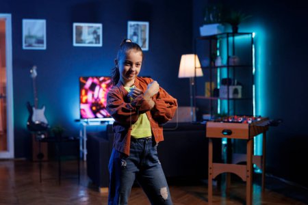 Foto de Linda chica que realiza la rutina de baile de tendencia en la sala de estar, rodeado de pantalla de TV que muestra renders 3D. Niño pequeño bailando dentro de las luces rgb iluminado estudio en casa, la creación de contenido para los fans en línea - Imagen libre de derechos