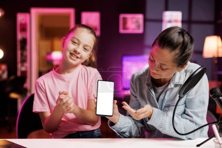 Glückliches Mädchen neben ihrer Mutter präsentiert eine Smartphone-Attrappe im rosafarbenen neonbeleuchteten Wohnzimmer, das als Vlogging-Studio dient. Junger Medienstar half Eltern, isoliertes Bildschirmtelefon zu präsentieren