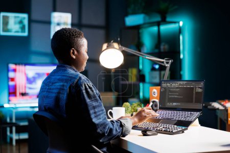 Afroamerikanische Freelancerin, die an einem Schreibtisch arbeitet, der sich auf ihren Laptop konzentriert, der Code und Daten anzeigt. Schwarze Frau mit drahtlosem Computer, programmiert und verwaltet die Software und sorgt für Cyber-Sicherheit.