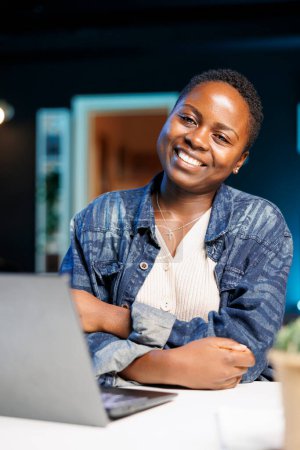 Foto de Retrato de una dama afroamericana mostrando una sonrisa genuina mientras está sentada frente a su computadora personal portátil. Mujer negra positiva sentada con los brazos cruzados cerca del portátil, mirando a la cámara. - Imagen libre de derechos