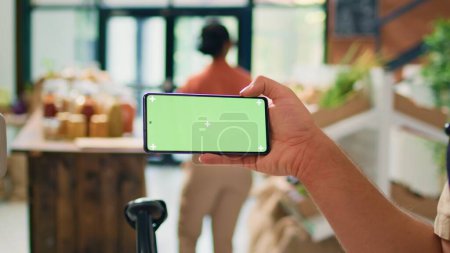 Verkäufer zeigt Greenscreen auf Telefondisplay, präsentiert isolierte Attrappe auf Gadget in lokalen Bio-Zero-Abfalllager. Junger Mann Verkäufer mit leerem Chromakey Copyspace-Bildschirm.