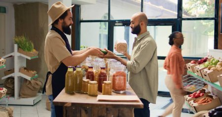 Shopper presents seasonings and products in glass jars to vegan customer while he is shopping for groceries. Middle eastern man wants to buy organic natural fresh fruits and veggies.