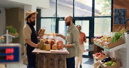 Founder of organic bio supermarket opens reusable packaging with bulk products, presents variety of pasta to shopper. Salesperson in apron suggests groceries free of synthetic chemicals.