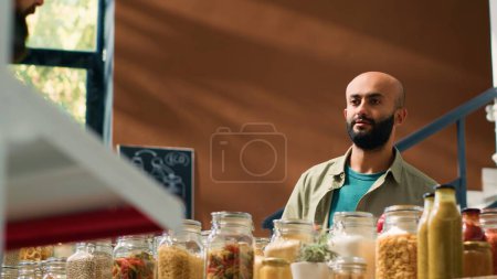 Photo for Seller offers man bulk products, presenting middle eastern buyer selection of pasta, herbs and spices. Young adult buying organic produce at nearby eco friendly supermarket. Handheld shot. - Royalty Free Image