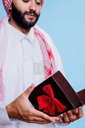 Photo for Muslim man wearing traditional cultural attire opening gift box for special occasion. Arab dressed in white thobe holding festive present giftbox with bow ribbon in hands closeup - Royalty Free Image