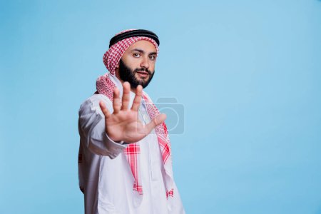 Foto de Hombre musulmán vistiendo ropa tradicional mostrando gesto de alto con la mano y mirando a la cámara con expresión seria. Arab confiado posando con prohibición signo estudio retrato - Imagen libre de derechos