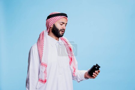 Téléchargez les photos : Homme musulman vêtu de vêtements culturels tenant des médicaments et lisant les instructions sur la bouteille. Personne portant une robe arabe traditionnelle et un foulard vérifiant l'emballage de vitamines - en image libre de droit