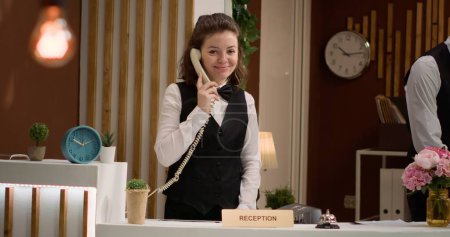 Front desk staff makes room reservations at hotel reception, using landline phone call to talk to guests about accommodation and bookings. Woman receptionist answering telephone. Handheld shot.