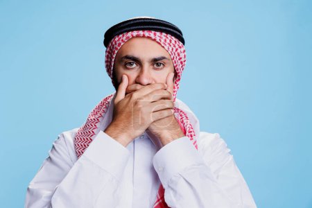 Man wearing traditional islamic clothes covering mouth with hands, showing speak no evil sign studio portrait. Muslim person keeping secret, showcasing three wise monkeys concept