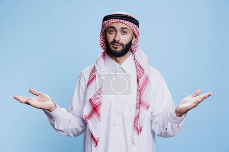 Photo for Pensive man wearing muslim thobe and headscarf, standing and shrugging shoulders while looking at camera. Arab person showing hesitation with open arms gesture studio portrait - Royalty Free Image