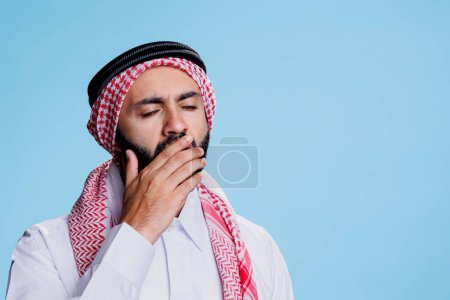 Téléchargez les photos : Homme musulman épuisé portant un foulard traditionnel et thobe bâillant et couvrant la bouche à la main. Personne fatiguée vêtue d'une coiffure et d'une robe de fantôme islamique faisant un geste somnolent - en image libre de droit