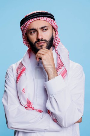 Hombre musulmán con atuendo tradicional contemplando con mirada pensativa. Interrogado árabe persona con thobe y ghutra pañuelo en la cabeza posando en pose de pensamiento con la mano apoyo barbilla