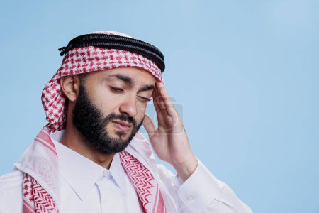Muslim man in traditional clothes expressing discomfort and rubbing temple while suffering from migraine attack. Arab person wearing ghutra headdress having headache and feeling pain