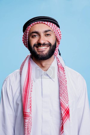 Foto de Hombre alegre musulmán vestido con toba blanca islámica y retrato de estudio de ghutra a cuadros. Persona árabe feliz de pie y mirando a la cámara con expresión despreocupada - Imagen libre de derechos