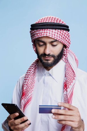 Man in traditional muslim clothes looking at credit card while making electronic payment on smartphone online store. Arab wearing ghutra and thobe processing purchase in internet shop