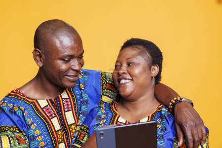 Téléchargez les photos : Joyeux couple noir souriant portant des vêtements ethniques étreignant tout en appréciant l'activité en ligne et en utilisant une tablette numérique. Heureux couple dans la relation embrassant et défilant les médias sociaux nourrir ensemble - en image libre de droit