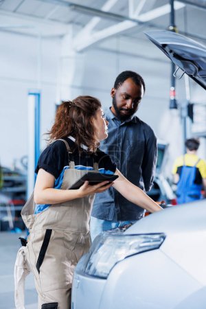 Mechanic in car service ordering new ignition system for damaged vehicle using tablet. Employee next to BIPOC client looking online for components to replace old ones in malfunctioning automobile