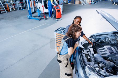 Photo for Skilled technician in garage finishing fixing car for BIPOC woman, looking underneath vehicle hood to remove remaining oil leaks. Worker does routine ignition system cleaning on client automobile - Royalty Free Image
