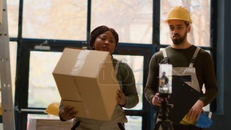 Photo for Warehouse employees using smartphone placed on tripod to make video for social media promoting the company. Coworkers filming themselves in storage room showcasing their daily tasks - Royalty Free Image
